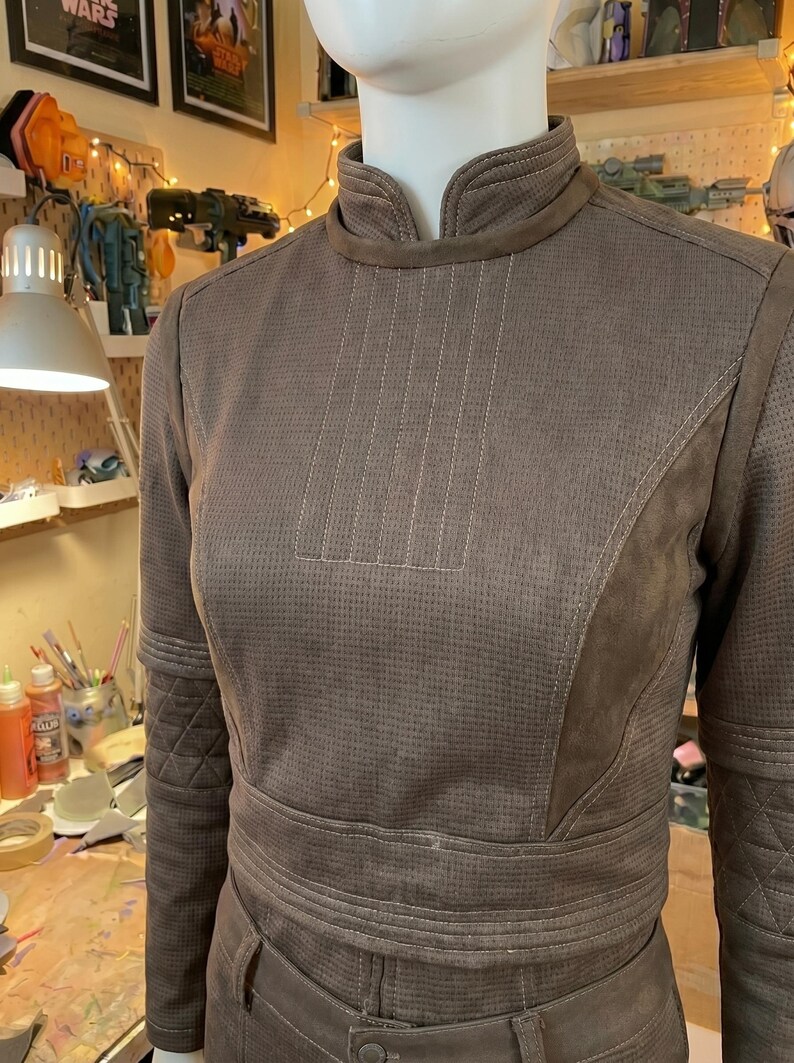 May include: A brown long-sleeved top with a high collar, featuring a textured micro-perforated fabric, vertical stitched detailing on the chest, and quilted diamond patterns on the upper sleeves, worn on a white mannequin torso.