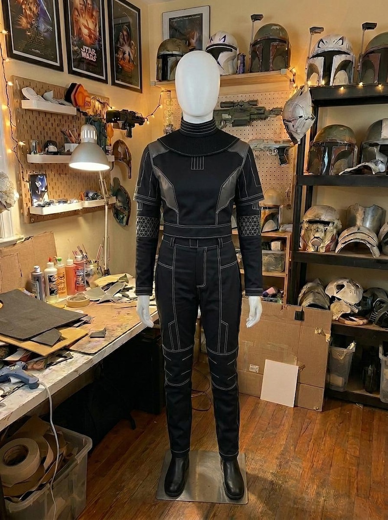 Puede incluir: Un mono t&aacute;ctico negro de manga larga con cuello alto, paneles estructurados en los hombros y costuras blancas geom&eacute;tricas. La prenda incluye un patr&oacute;n de diamantes texturizado en los antebrazos y botas negras a juego.