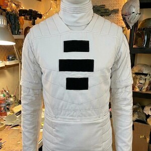 Puede incluir: Una camisa blanca de manga larga con cuello alto y patrones de costura acolchada horizontal. El pecho presenta tres parches rectangulares negros dispuestos verticalmente en el centro. La prenda se muestra en un maniqu&iacute;.