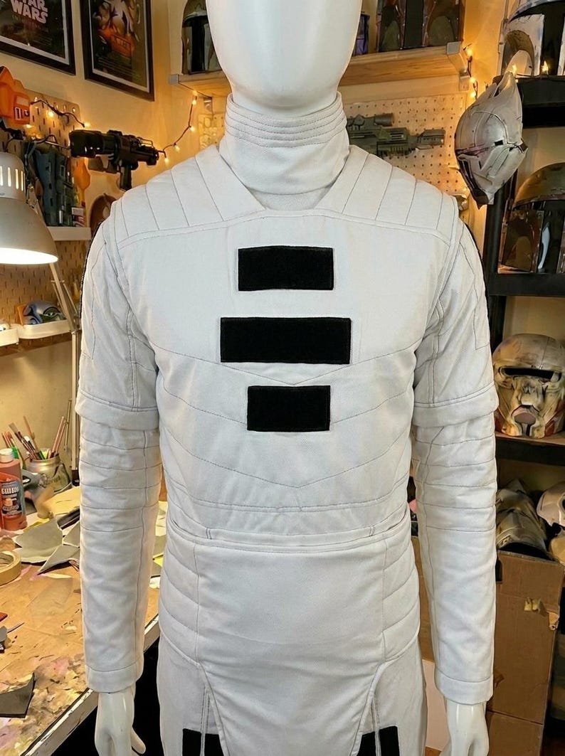 Puede incluir: Una camisa blanca de manga larga con cuello alto y patrones de costura acolchada horizontal. El pecho presenta tres parches rectangulares negros dispuestos verticalmente en el centro. La prenda se muestra en un maniqu&iacute;.