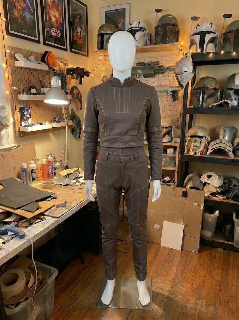 May include: A brown two-piece outfit consisting of a long-sleeved cropped top with a high collar and quilted elbow patches, paired with matching trousers featuring a button closure, belt loops, and reinforced knee panels with visible stitching.