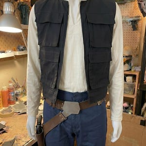 May include: A costume ensemble featuring a cream-colored long-sleeve shirt with a mandarin collar, a black utility vest with four front pockets, dark blue trousers, and a brown leather-style belt with a metal buckle and a side holster.