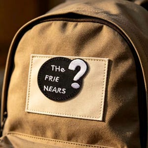 May include: A tan backpack featuring a rectangular beige patch on the front. Centered on the patch is a circular black embroidered patch with white text that reads "THe FRIE NEARS" and a large white question mark symbol to the right side.