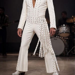 May include: A white jumpsuit featuring a deep V-neckline, long sleeves, and flared pant legs. The garment is adorned with silver-toned metal studs along the front, the waist belt, and the long fringe strips hanging from the right side of the waist.