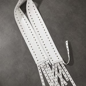 May include: A white belt featuring two parallel rows of silver-toned metal grommets running the entire length of the strap. The end of the belt is divided into multiple long, narrow strips, each finished with a single silver-toned metal grommet.