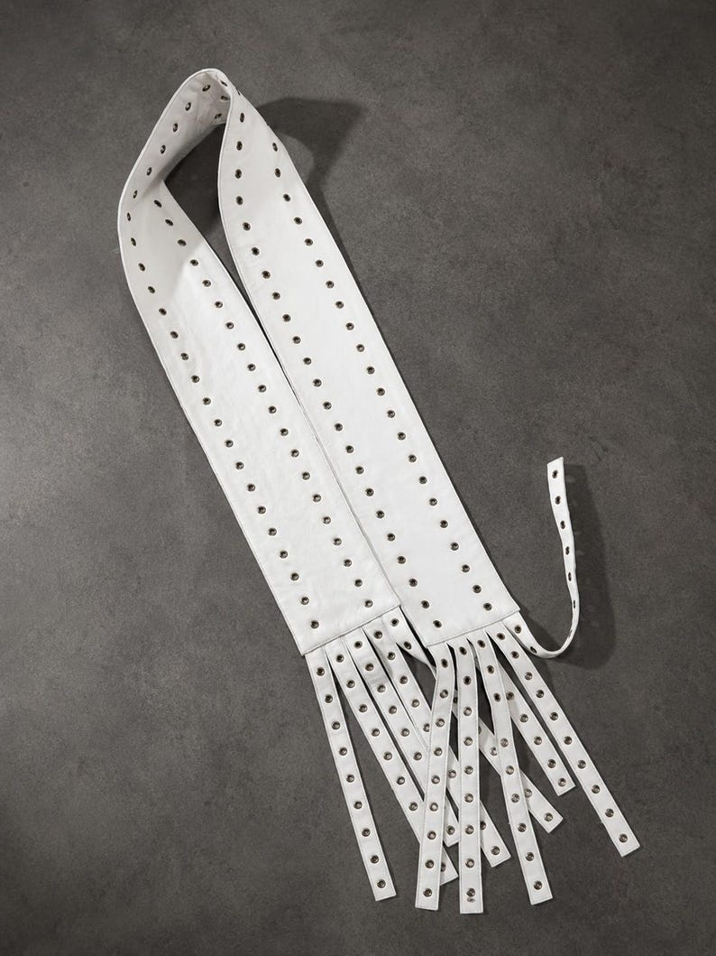 May include: A white belt featuring two parallel rows of silver-toned metal grommets running the entire length of the strap. The end of the belt is divided into multiple long, narrow strips, each finished with a single silver-toned metal grommet.