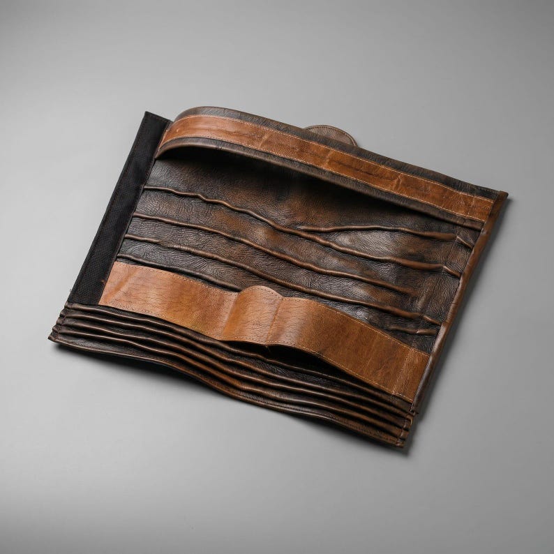 May include: A brown leather tool roll featuring multiple horizontal pockets for storage. The organizer has a dark brown textured finish with lighter tan leather accents along the top and bottom edges, designed to fold and secure various small items.