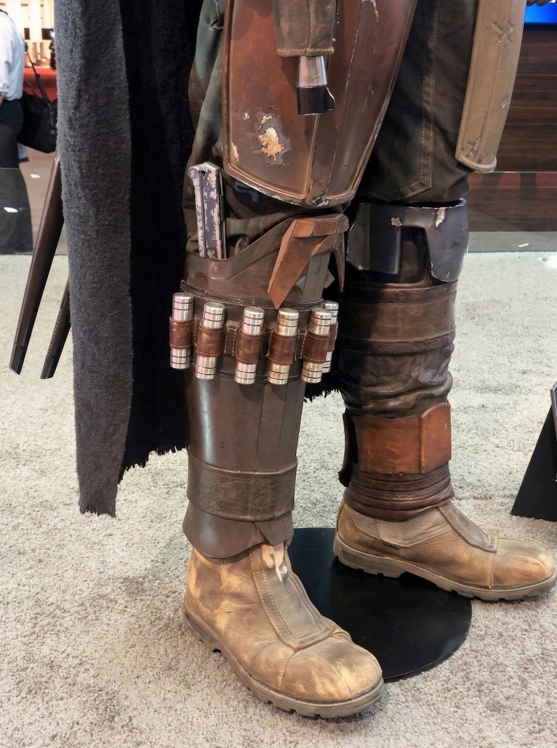 May include: A costume featuring brown leather-like leg armor with a row of six metallic cylindrical canisters attached to the shin. The armor is worn over dark trousers and tan, weathered-looking boots. A dark, textured cape hangs to the side.