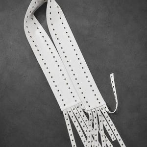 May include: A long white belt featuring two parallel rows of metal grommets running the entire length. The end of the belt is cut into multiple thin, dangling strips, each finished with a single metal grommet at the tip, creating a fringe effect.