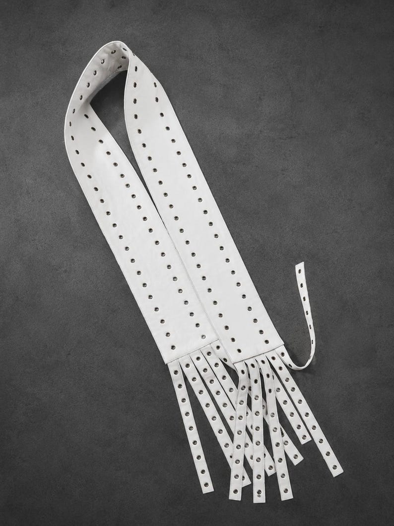 May include: A long white belt featuring two parallel rows of metal grommets running the entire length. The end of the belt is cut into multiple thin, dangling strips, each finished with a single metal grommet at the tip, creating a fringe effect.
