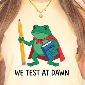 May include: A pale yellow t-shirt featuring a graphic of a green frog wearing a red cape, holding a yellow pencil and a blue book labelled Calculus. Below the frog, the text reads WE TEST AT DAWN in bold black capital letters.