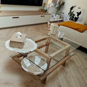 Peut inclure: Table basse carrée en bois avec plateau en verre. Sur la table se trouvent une boîte à mouchoirs tissée, un dessous de plat rond tissé et un bol décoratif avec des sections tissées blanches et naturelles. La pièce a une palette de couleurs neutres.