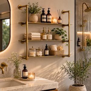 May include: A three-tier wall-mounted shelf unit constructed from gold-toned metal pipes and flat shelves. The shelves hold various bathroom items including amber glass bottles, folded beige towels, a woven basket, and potted green plants.