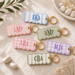 May include: Six rectangular scalloped keychains with gold-tone metal rings. The keychains feature embroidered monograms: EKA on blue stripes, LMP on pink, HMP on sage green, LRA on pink gingham, MJS on purple gingham, and CBA on green stripes.