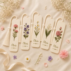 May include: Five rectangular off-white bookmarks with rounded corners, each featuring a unique embroidered floral design, a hanging tassel, and the name Olivia printed in cursive script. The flowers include pink carnations, purple bellflowers, white daisies, lily of the valley, and a pink rose.