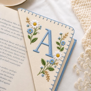 May include: A triangular felt corner bookmark in cream featuring a blue embroidered letter A, surrounded by detailed floral motifs in white, blue, and green. The edges are finished with blue blanket stitching, securing the bookmark to a book page.