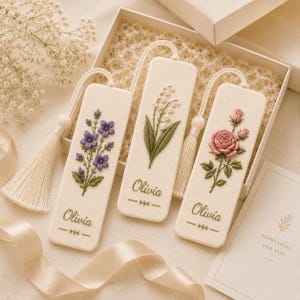 May include: Three rectangular off-white bookmarks with cream-colored tassels, each featuring embroidered floral designs of purple bellflowers, lily of the valley, and a pink rose. The name Olivia is embroidered in green on each bookmark.