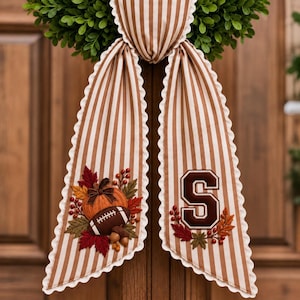 May include: A decorative fabric bow with brown and white vertical stripes and scalloped edges. One tail features an embroidered pumpkin, football, and autumn leaves. The other tail displays a large, dark brown embroidered letter S with leaves.