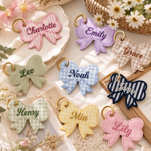 May include: A collection of nine fabric bow keychains in various colors and patterns, each embroidered with a unique name including Charlotte, Emily, Grace, Leo, Noah, James, Henry, Mia, and Lily. Each bow features a gold-toned metal keyring.