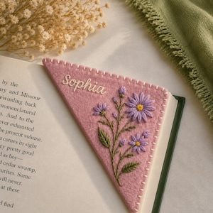 May include: A triangular felt bookmark in a dusty pink shade, featuring hand-stitched edges in white thread. The surface displays an embroidered floral arrangement of purple daisies with yellow centers and green stems, alongside the name Sophia in white.