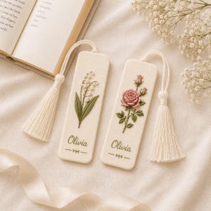 May include: Two rectangular off-white fabric bookmarks featuring embroidered floral designs. One displays lily of the valley, the other a pink rose. Both are personalized with the name Olivia in green script, include a cream tassel, and have a decorative line motif.
