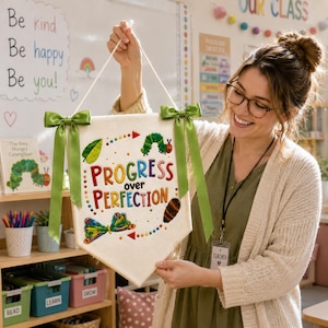 May include: A cream-colored fabric wall banner featuring the embroidered text "PROGRESS over PERFECTION" in colorful letters. The design includes a green leaf, a caterpillar, a butterfly, and a chrysalis, with green satin ribbons tied at the top corners.