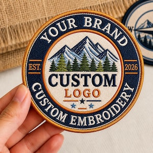 May include: A circular embroidered patch featuring a mountain range and pine forest graphic. The text reads YOUR BRAND, EST. 2026, CUSTOM LOGO, and CUSTOM EMBROIDERY. The design uses navy blue, cream, green, and orange thread with a gold border.