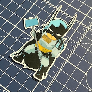 May include: A sticker of a cartoon Batman character, holding a blue hammer. The character is in black and light blue, with a yellow utility belt. The sticker is on a blue grid background. The image has a comic book style.