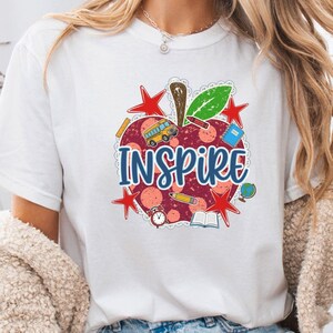May include: A white short-sleeve t-shirt featuring a large graphic of a red apple with the word INSPIRE in blue letters. The apple design includes small illustrations of a school bus, pencil, globe, book, alarm clock, and red stars.