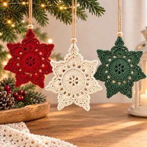 Puede incluir: Tres adornos en forma de estrella hechos de hilo de ganchillo en rojo, crema y verde oscuro. Cada adorno presenta un patrón circular central con puntas radiantes, suspendido por un cordel de yute con una pequeña cuenta de madera arriba.