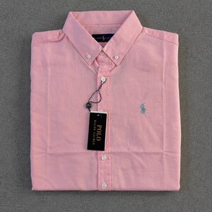 Può includere: Una camicia rosa chiaro con bottoni, colletto classico e maniche lunghe. La camicia presenta un piccolo logo blu sul petto e un'etichetta nera con la scritta "POLO RALPH LAUREN". La camicia è ben piegata.