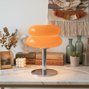 Peut inclure: Une lampe de table orange au design rétro, avec un abat-jour arrondi superposé et une base chromée. La lampe est posée sur une surface en marbre blanc, avec des livres, des vases et une petite image encadrée en arrière-plan. Le cordon de la lampe est visible.