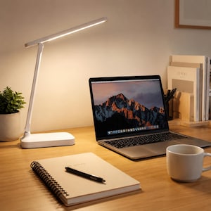 Peut inclure: Une lampe de bureau LED blanche avec un bras fin réglable et une base rectangulaire, émettant une lumière chaude. La lampe est posée sur une surface en bois à côté d'un ordinateur portable ouvert, d'un carnet à spirales et d'une tasse blanche.