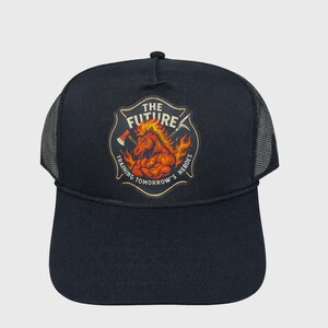 May include: Black trucker hat with a mesh back and a curved brim. The front features a shield-shaped emblem with a horse engulfed in flames, axes, and the text "THE FUTURE TRAINING TOMORROW'S HEROES".