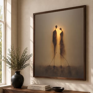 Könnte beinhalten: Ein quadratisches Wandbild in einem dunklen Holzrahmen zeigt die Silhouette zweier Figuren, die vor einem sanften, goldenen Licht stehen. Das Kunstwerk hat eine dunstige Ästhetik mit dunklen, länglichen Formen auf einem neutralen Hintergrund.