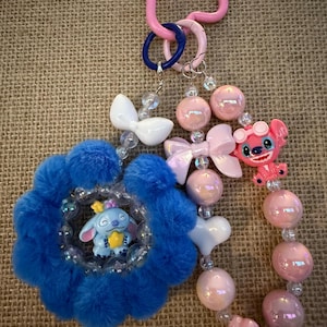 May include: A handmade keychain featuring a blue flower-shaped charm with a Stitch figurine, pink and white beads, and a pink carabiner clip. The keychain includes a pink Stitch figurine with sunglasses.