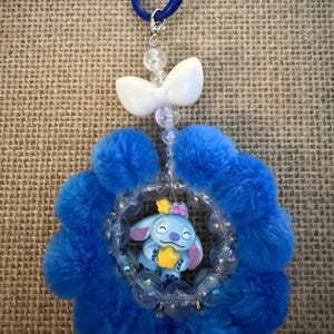 May include: A blue and white charm featuring a cartoon character holding a star, surrounded by a fluffy blue flower shape. The charm is attached to a blue ring with a white bow and clear beads.