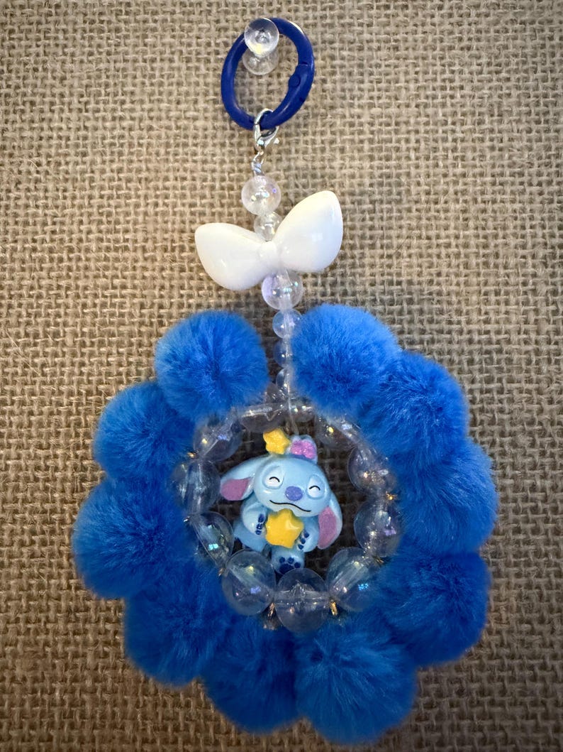 May include: A blue and white charm featuring a cartoon character holding a star, surrounded by a fluffy blue flower shape. The charm is attached to a blue ring with a white bow and clear beads.