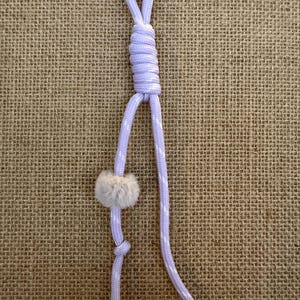 May include: A light purple cord with white polka dots, knotted and looped at the top. A small, fluffy, gray pom-pom is attached to the cord. The cord is displayed against a textured, tan background.