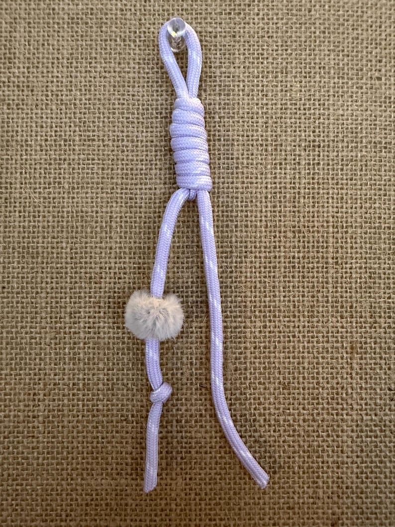 May include: A light purple cord with white polka dots, knotted and looped at the top. A small, fluffy, gray pom-pom is attached to the cord. The cord is displayed against a textured, tan background.