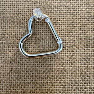May include: A silver-colored heart-shaped carabiner with a clear plastic stud. The carabiner is made of metal and has a spring-loaded gate. The background is a textured, tan fabric.