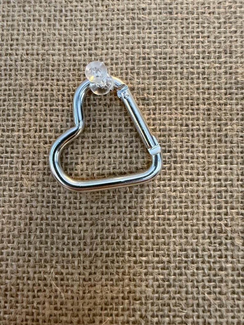 May include: A silver-colored heart-shaped carabiner with a clear plastic stud. The carabiner is made of metal and has a spring-loaded gate. The background is a textured, tan fabric.