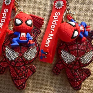 Op de afbeelding: Twee Spider-Man sleutelhangers met een rood en blauw siliconen figuur bevestigd aan een rode band met de tekst Spider-Man. Elke sleutelhanger bevat een rood, met strass steentjes ingelegd Spider-Man masker met witte kristallen ogen.