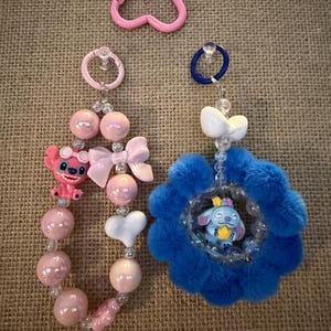 May include: Pink and blue beaded keychains with character charms. One keychain features a pink Stitch figure, pink beads, and a white bow. The other has a blue flower design with a Stitch figure inside. A pink heart-shaped carabiner is also visible.