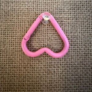 May include: A pink heart-shaped carabiner clip with a clear suction cup attached. The clip is made of a solid, smooth material and has a small hinge on one side. The background is a textured, tan fabric.