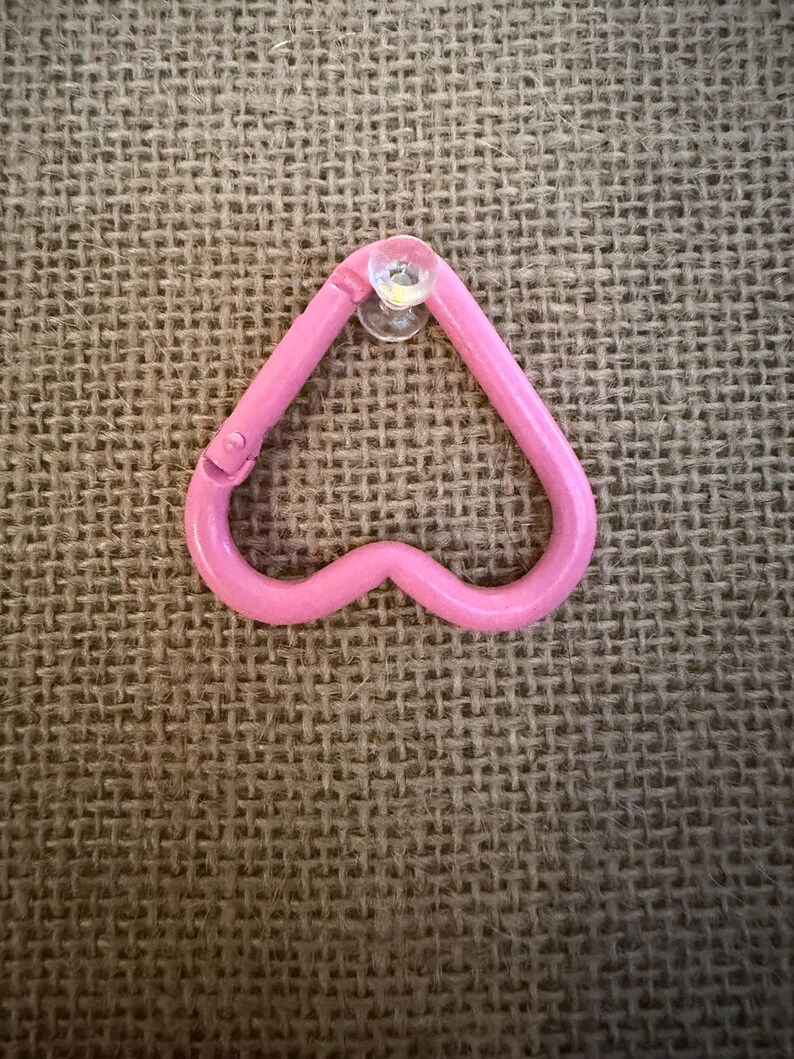 May include: A pink heart-shaped carabiner clip with a clear suction cup attached. The clip is made of a solid, smooth material and has a small hinge on one side. The background is a textured, tan fabric.