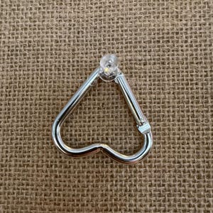 May include: A silver-colored heart-shaped carabiner clip with a clear, rounded top. The clip is made of metal and has a spring-loaded gate for secure closure. The background is a textured, tan fabric.