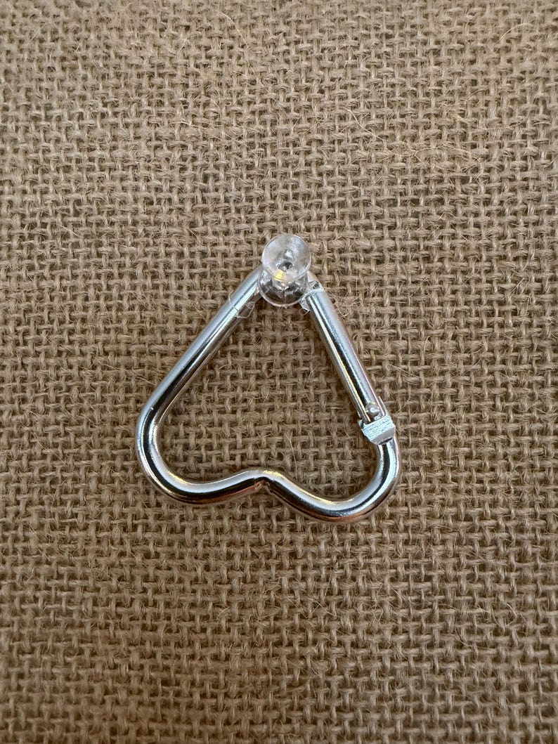 May include: A silver-colored heart-shaped carabiner clip with a clear, rounded top. The clip is made of metal and has a spring-loaded gate for secure closure. The background is a textured, tan fabric.