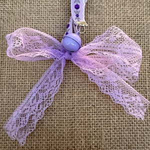 May include: A lavender lace bow with a matching ribbon and a small bell. The ribbon has a decorative pattern with purple accents. The bow is made of delicate, open-weave lace.