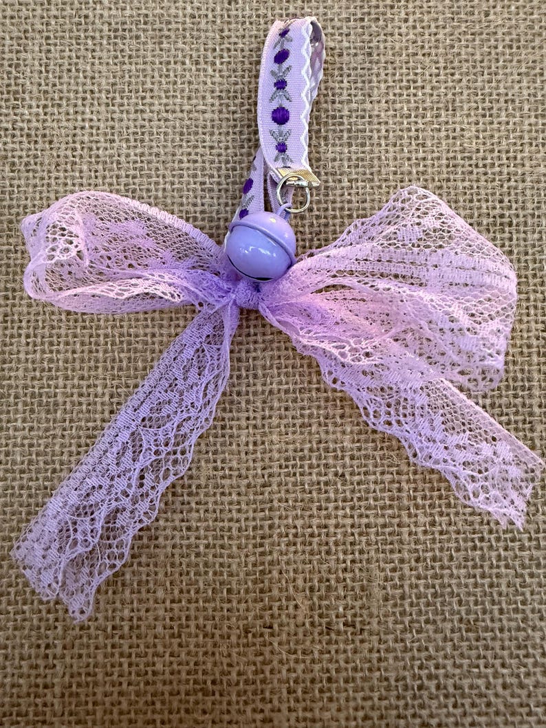 May include: A lavender lace bow with a matching ribbon and a small bell. The ribbon has a decorative pattern with purple accents. The bow is made of delicate, open-weave lace.
